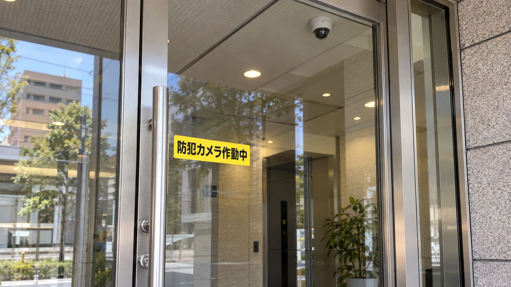事務所のガラスの入口扉の目の高さに貼った防犯カメラ作動中のステッカーと、扉の上の防犯カメラ